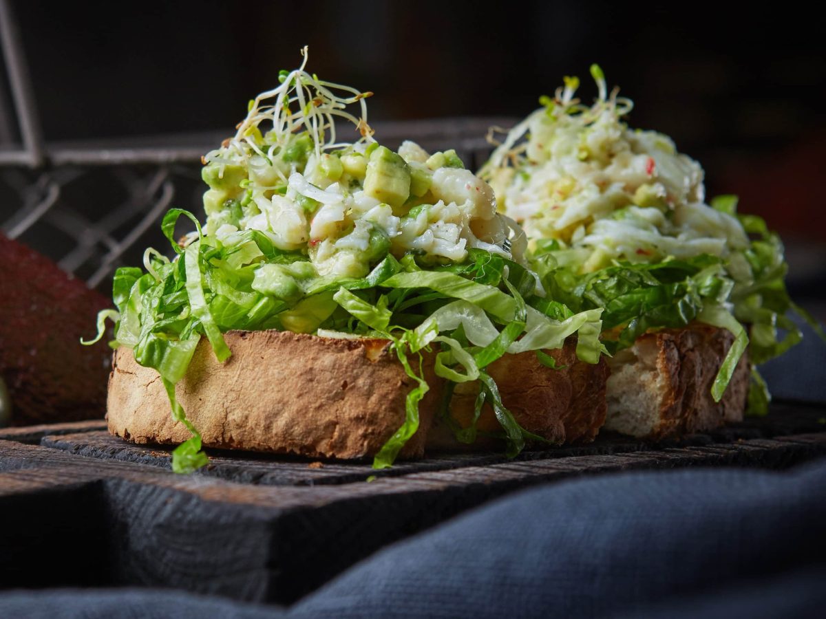 Ein dunkler Hinter-und Untergrund. In der Mitte das Sandwich mit dem Avocado-Garnelen-Eiersalat seitlich fotografiert.