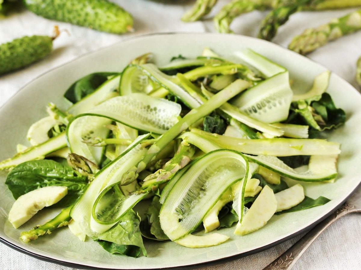 Ein Teller Spargel-Gurken-Salat, mit Babyspinat, daneben eine Gabel und Gurken sowie Spargelstangen.