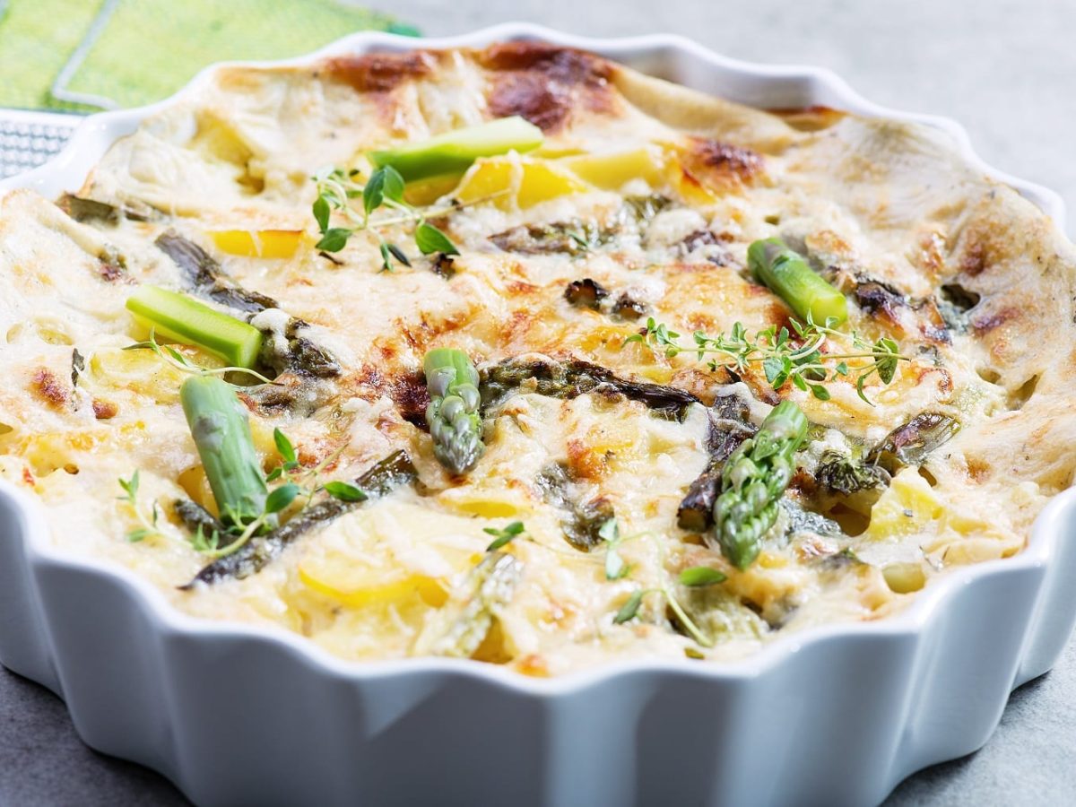 Eine weiße Auflaufform mit Kartoffel-Spargelgratin auf einem grauen Tisch. Hinten links unscharf ein grünes Tuch mit einer Metallreibe zu erkennen.