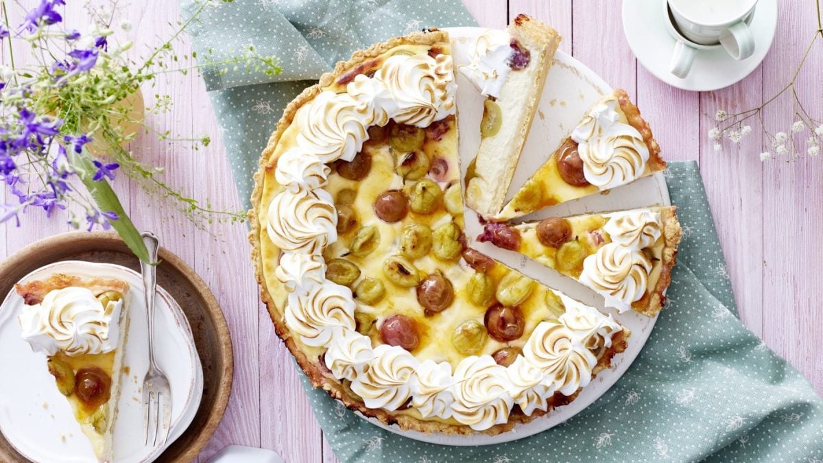 Ein Käsekuchen mit Baiser und Stachelbeeren angeschnitten auf einer weißen Kuchenplatte auf einem grünem Geschirrtuch mit weißen Punkten. Links oben Blumen und links unten ein Stück Kuchen auf einem Teller. Rechts oben Espressotassen.