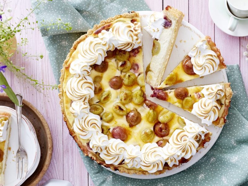 Ein Käsekuchen mit Baiser und Stachelbeeren angeschnitten auf einer weißen Kuchenplatte auf einem grünem Geschirrtuch mit weißen Punkten. Links oben Blumen und links unten ein Stück Kuchen auf einem Teller. Rechts oben Espressotassen.