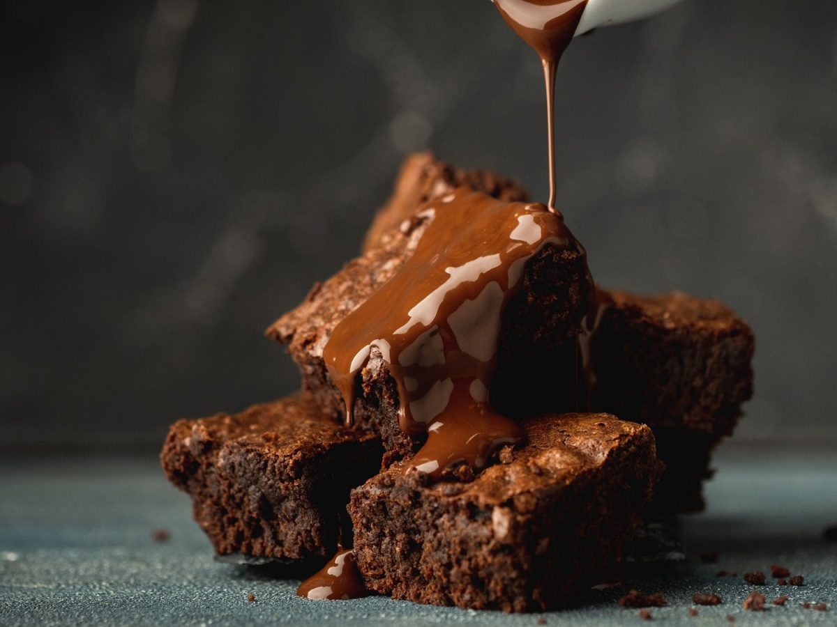 Ein paar Tahini-Brownies auf ebenem Untergrund, die mit flüssiger Schokolade übergossen werden.