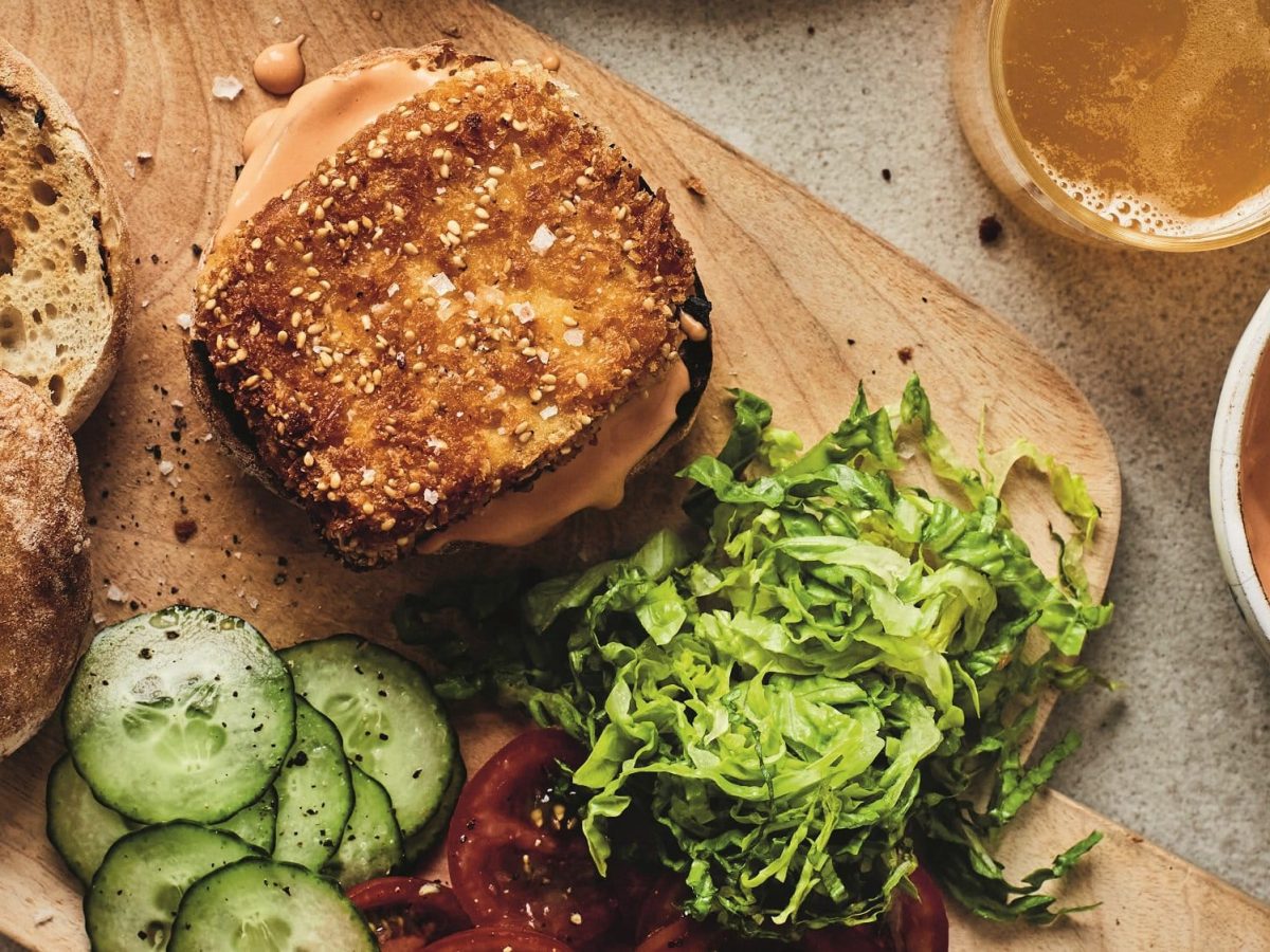 Tofu-Burger auf einem Brett, auf dem frische Tomaten, Gurken und Salat geschnitten liegen. Alles Draufsicht.