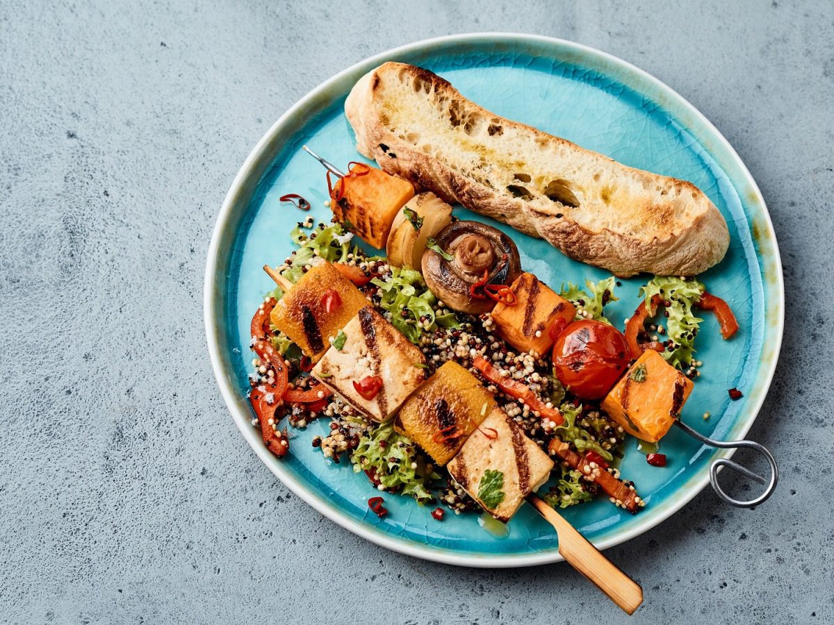 Zwei gegrillte Tofu-Gemüsespieße auf einem Teller, daneben eine Scheibe Baguette.