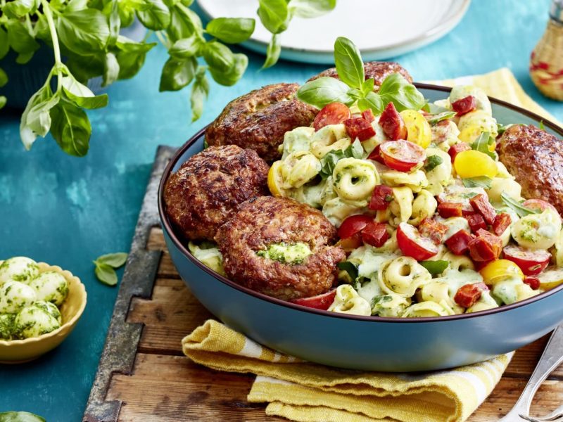 Eine blaue Bowl mit Tortellinisalat und Hackbällchen auf einem gelben Geschirrtuch auf einem hölzernen Brett. Der Hintergrund ist blau und links eine kleine Schüssel mit Mozzarellakugeln und ein Basilikumstrauch. Rechts oben ein Salzstreuer.