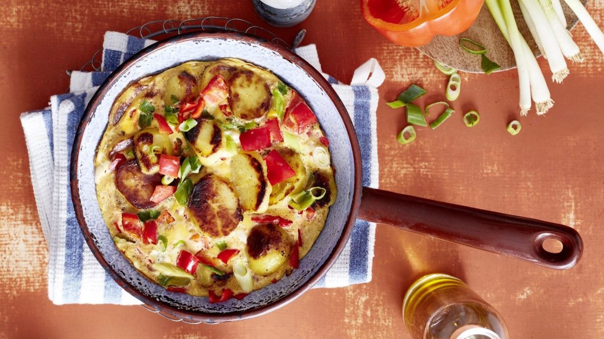 Eine blaue Pfanne mit braunen Griff, gefüllt mit einem Tortilla mit Paprika, auf einem blau/weiß gestreiften Geschirrtuch auf einem orangen Hintergrund. Rechts oben Tomaten und Lauchzwiebeln.