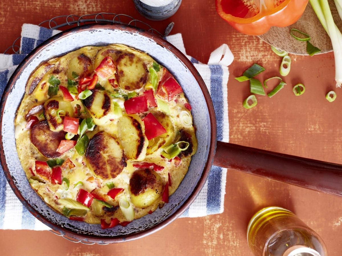 Eine blaue Pfanne mit braunen Griff, gefüllt mit einem Tortilla mit Paprika, auf einem blau/weiß gestreiften Geschirrtuch auf einem orangen Hintergrund. Rechts oben Tomaten und Lauchzwiebeln.