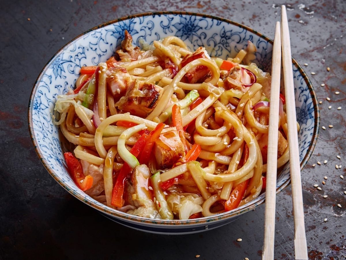 Eine weiß-blaue Schüssel auf dunklem Untergrund mit den Udon-Nudeln und zwei Essstäbchen auf der rechten Seite der Schüssel.