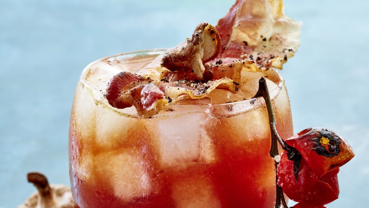 Umami-Tsunami im Glas vor hellblauem Hintergrund, garniert mit Tomaten, Bacon, Pilzen und Pfeffer.