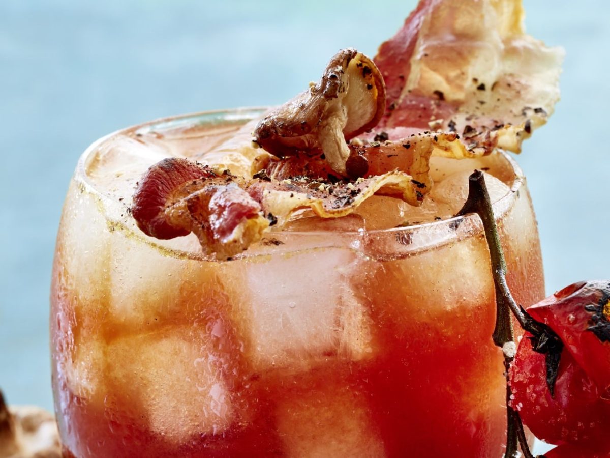 Umami-Tsunami im Glas vor hellblauem Hintergrund, garniert mit Tomaten, Bacon, Pilzen und Pfeffer.