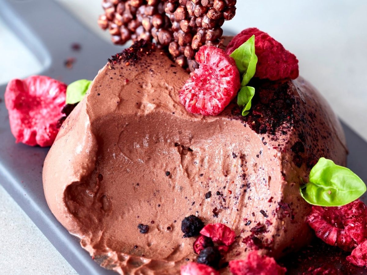 Eine Portion vegane Schokomousse mit getrockneten Himbeeren und Krokant und Basilikum auf einem Brettchen, heller Hintergrund.