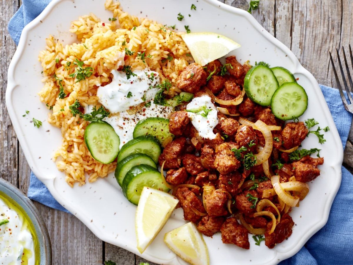 Eine weiße Platte mit vegetarischem Gyros, Reis, Zitronen und Gurken auf einem blauen Geschirrtuch auf einem hölzernen Hintergrund. Links unten ein Joghurt-Dip, rechts zwei Gabeln.