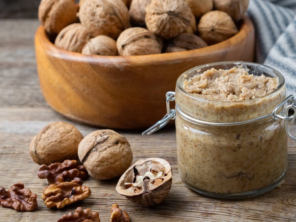 Frontal: Auf einem Untergrund aus Holz steht ein Glas Walnuss Pesto. Drum herum liegen ganze und aufgebrochene Walnüsse.