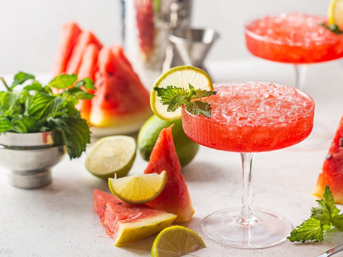 Zwei Gläser Wassermelonen-Daiquiri, darum frische Wassermelone und MInze sowie Cocktail-Zubehör.