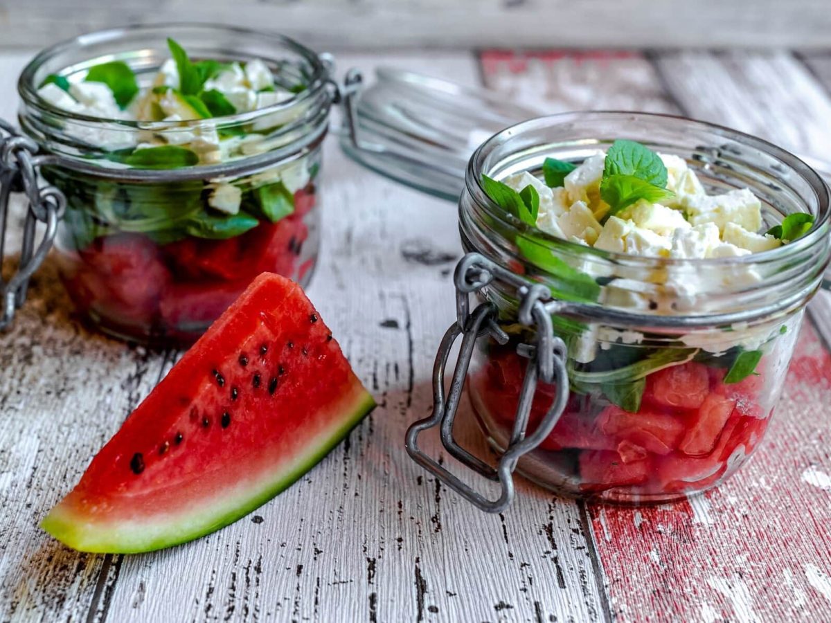 Frontal: Wassermelonensalat mit Feta und Minze im Glas, Schichtsalat auf hellem Holzboden.