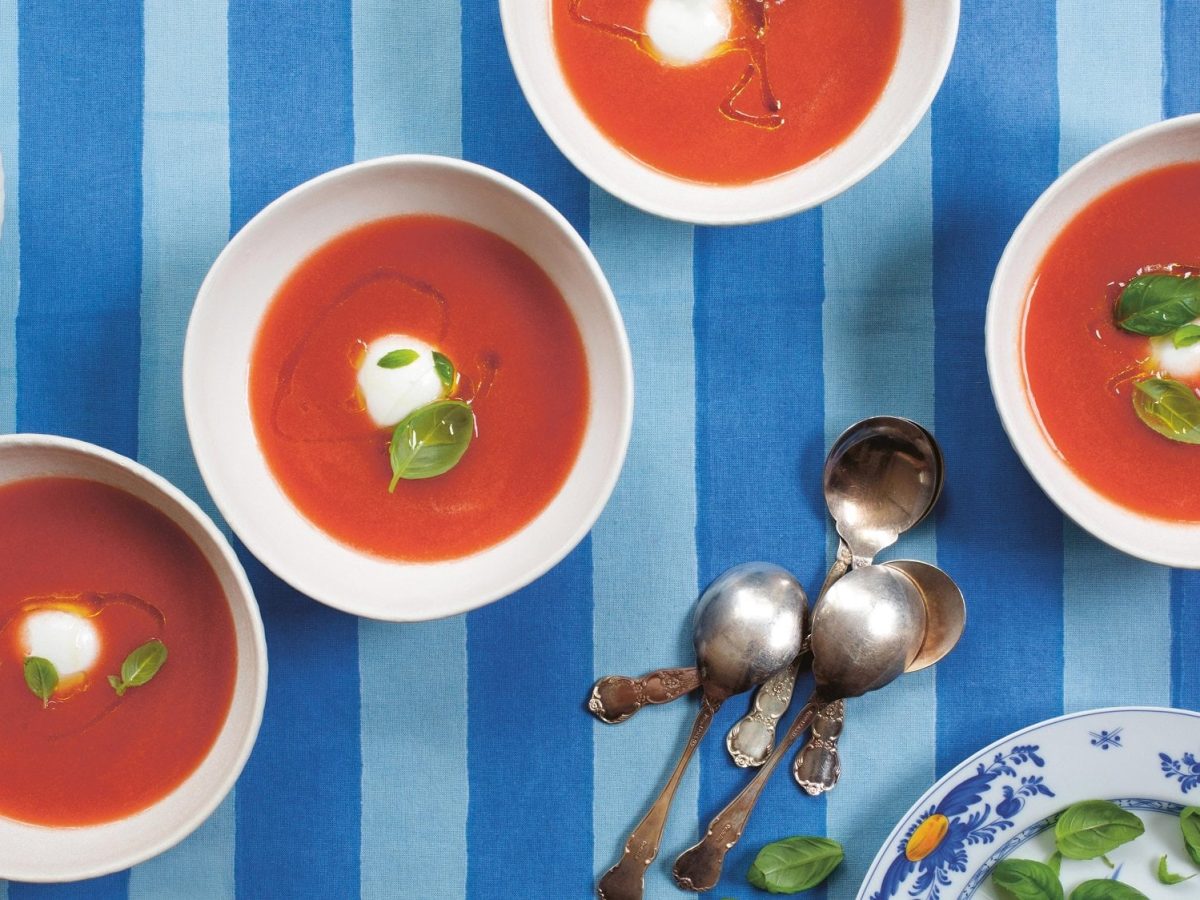 Ein paar weiße Schüsseln mit der Tomatensuppe Acgua de Pomodoro garniert mit Basilikum, Öl und Mozzarella, daneben drei Löffe, alles Draufsicht.