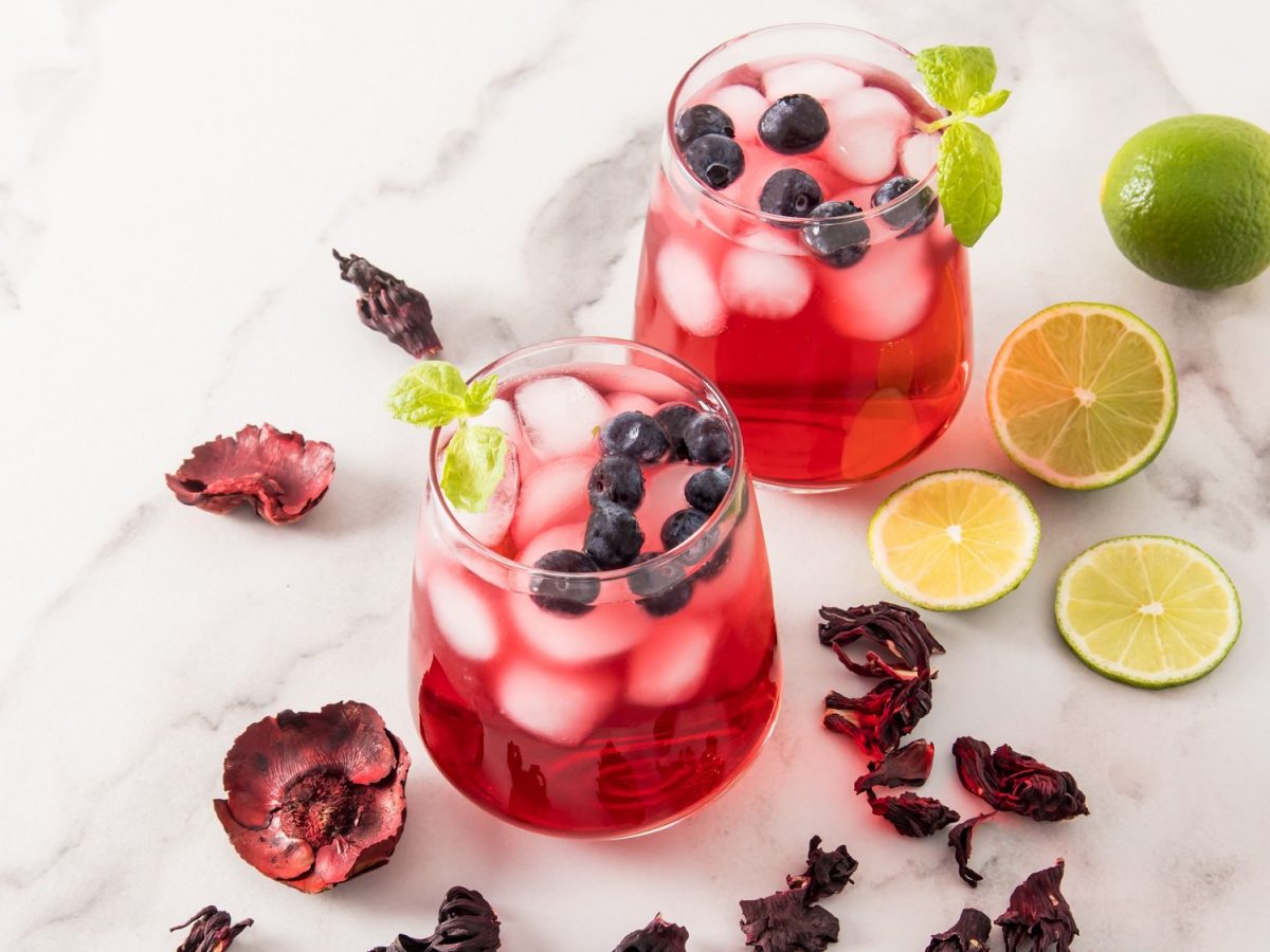 Zwei Gläser gefüllt mit dem Hibiskus-Eistee Agua de Jamaica, Eiswürfeln und Blaubeeren, drumherum getrocknete Hibiskusblüten, Limetten und Blaubeeren.