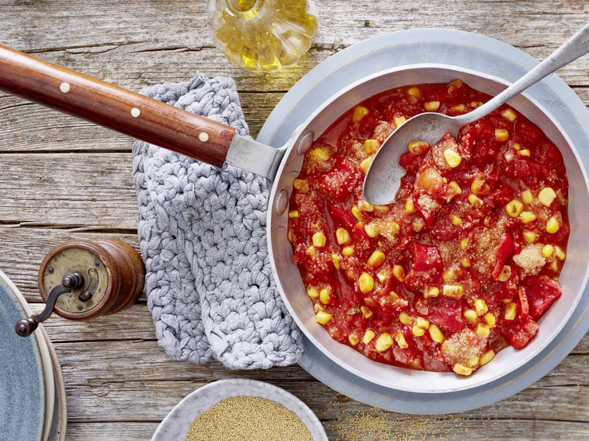 Eine Pfanne gefüllt mit Amaranth mit Paprika und Mais auf einem hölzernen Hintergrund. Links davon ein Topflappen, eine Pfeffermühle und Teller. Oben eine Flasche mit Öl und unten auf dem Bild eine kleine Schüssel mit Amaranth-Körnern.