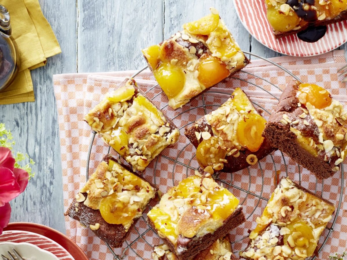 Acht Aprikosen-Schoko-Butterkuchen Stücke auf einem Kuchengitter auf einem orange/weißem Geschirrtuch. Links davon Blumen, eine Kaffeekanne und Teller. Rechts oben ein Teller mit einem Kuchenstück und einer Dessertgabel.