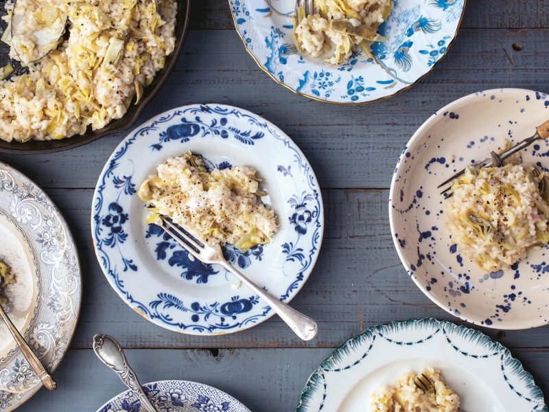 Mehrere blau-weiß-gemusterte Teller mit Artischocken-Risotto und Gabeln in der Draufsicht, außerdem eine große Pfanne mit mehr Risotto.