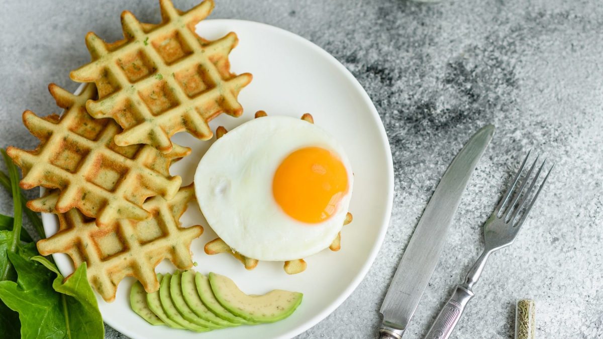 Knusprige Avocadowaffeln mit Spinat und Spiegelei auf einem weißen Teller und grauem Untergrund.