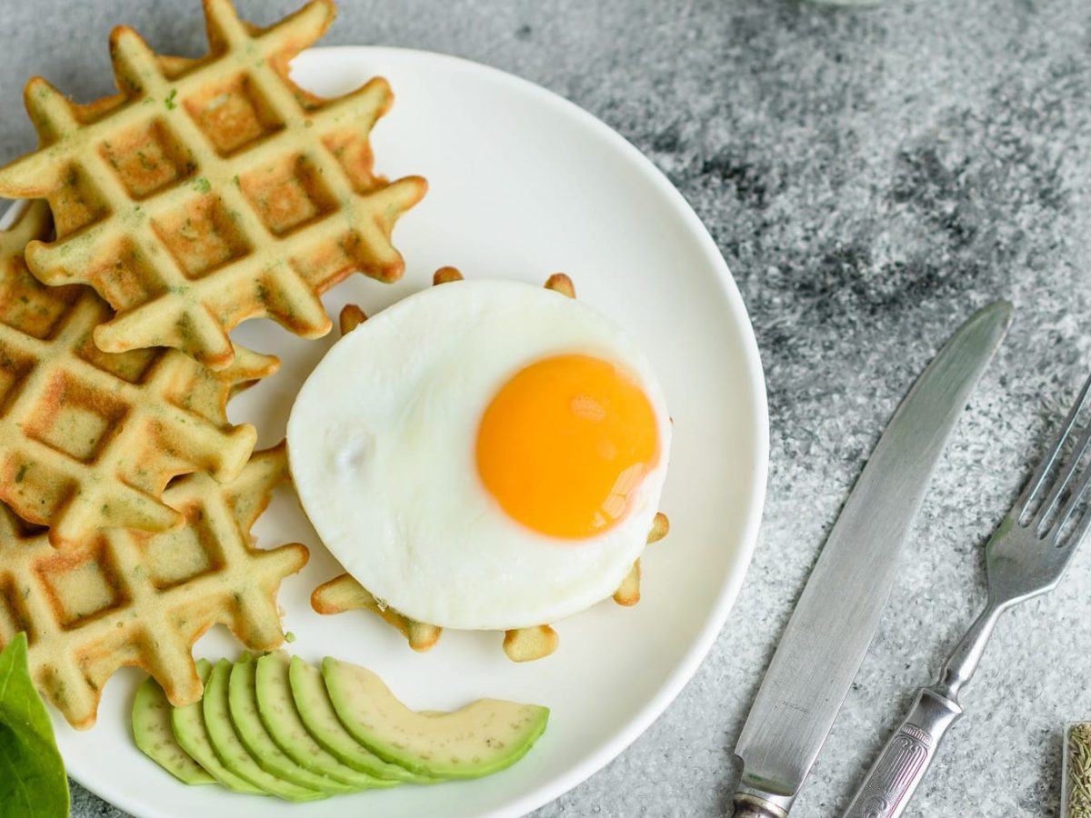 Knusprige Avocadowaffeln mit Spinat und Spiegelei auf einem weißen Teller und grauem Untergrund.