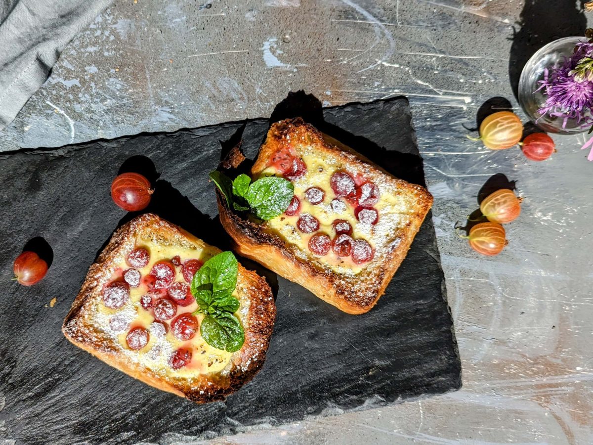 Zwei Brioche-Scheiben mit Quarkcreme und Stachelbeeren überbacken auf einer schwarzen Schieferplatte und grauem Untergrund von oben fotografiert.