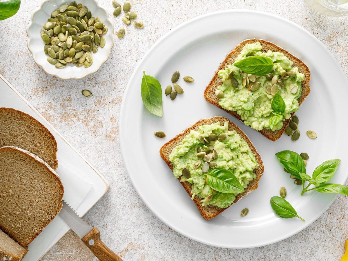 Zwei Brotscheiben mit Basilikum-Avocado, Kürbiskernen und Basilikumblättern auf einem Teller in der Draufsicht, drumherum ein angeschnittener Brotlaib, ein Stück Zitrone, eine Schüssel mit der Guacamole und Basilikum.