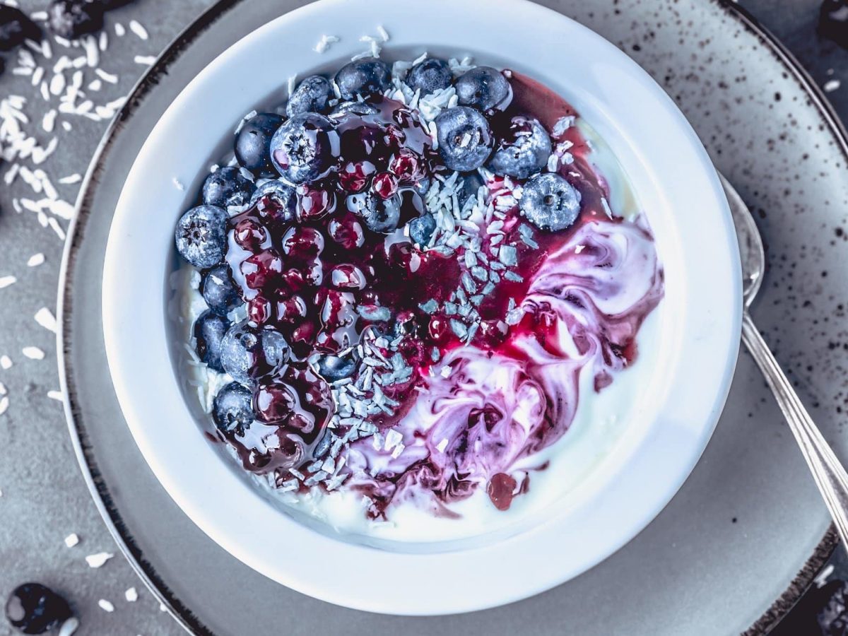 Draufsicht: eine weiße Schale Blaubeer-Kokos-Granola mit Quinoa steht auf einem hellem Teller, der auf dunklem Untergrund aus Holz steht.