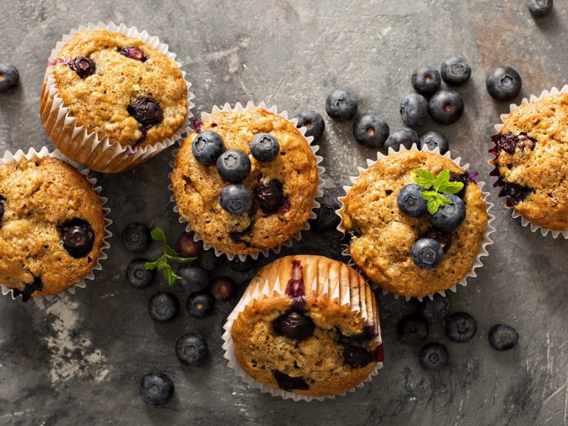 Mehrere Blaubeermuffins auf einem grauen Steintisch mit frischen Blaubeeren und Minze.