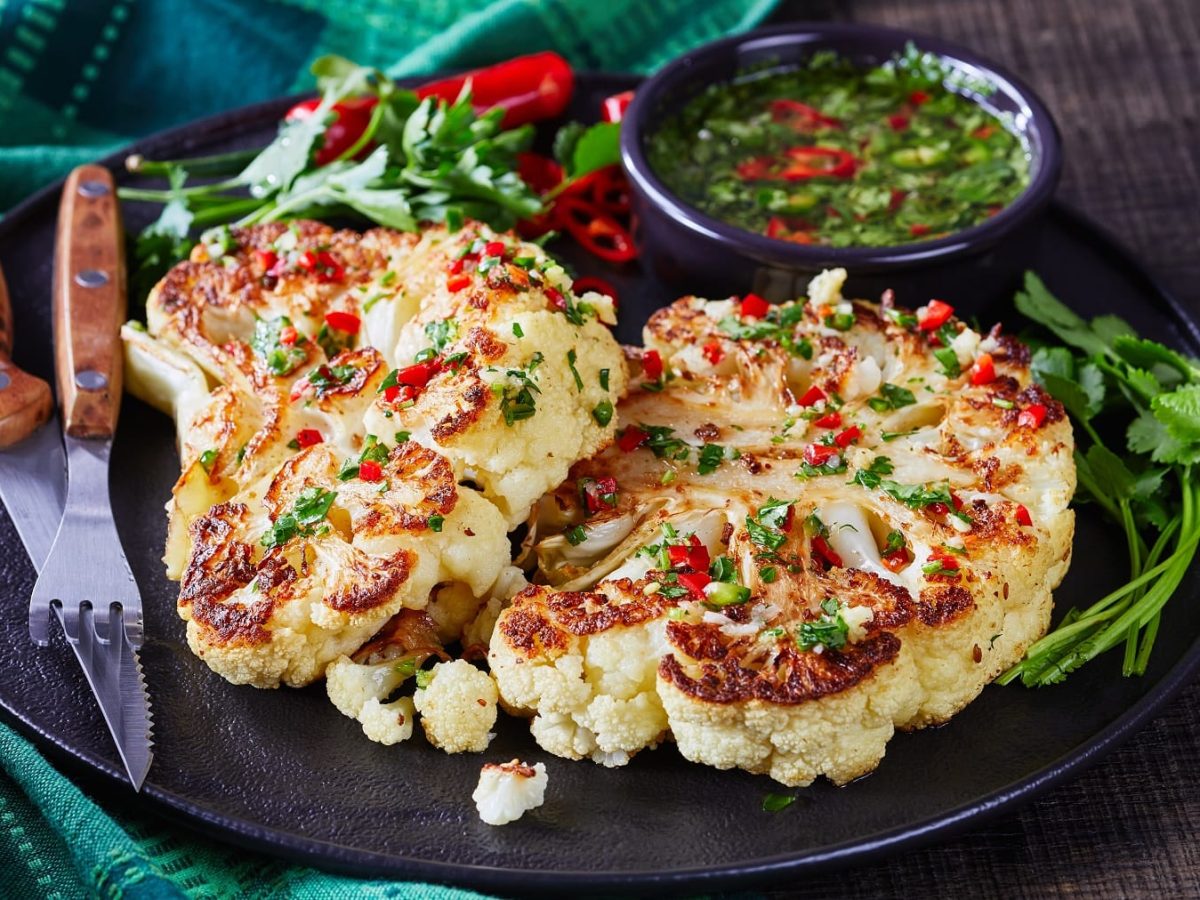 Zwei Blumenkohlsteaks auf einem schwarzen Teller mit asiatischem Dip und dunklem Untergrund. Daneben eine türkisfarbene Serviette.