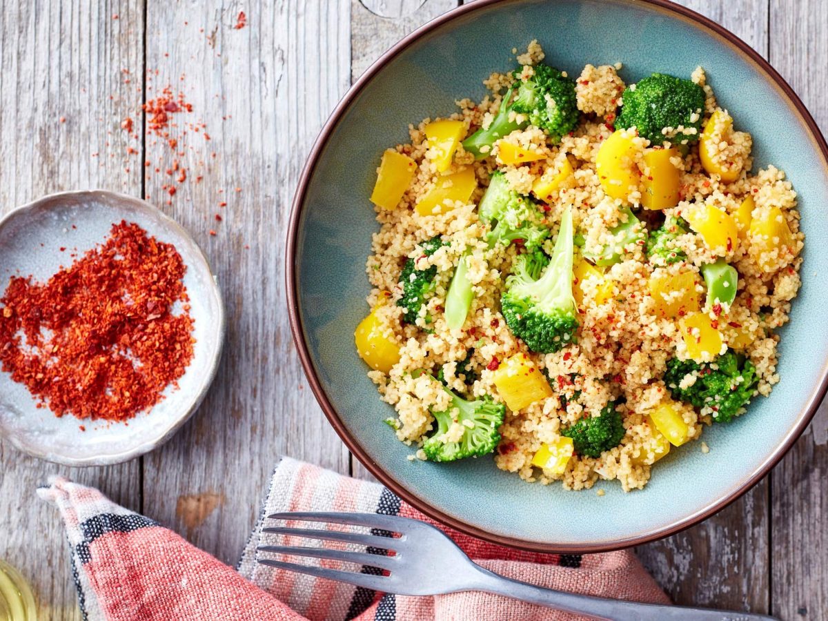 Eine Brokkoli-Bowl mit Couscous auf einem hölzernen Hintergrund. Links von der blauen Schüssel steht eine kleine Schüssel gefüllt mit Chiliflocken. Unten ein Geschirrtuch mit einer Gabel.