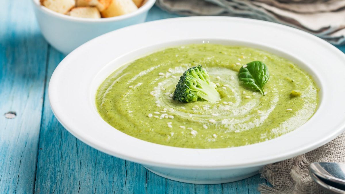 Ein weißer Teller mit der grünen Brokkoli-Cremesuppe auf einem bläulichen Holztisch.