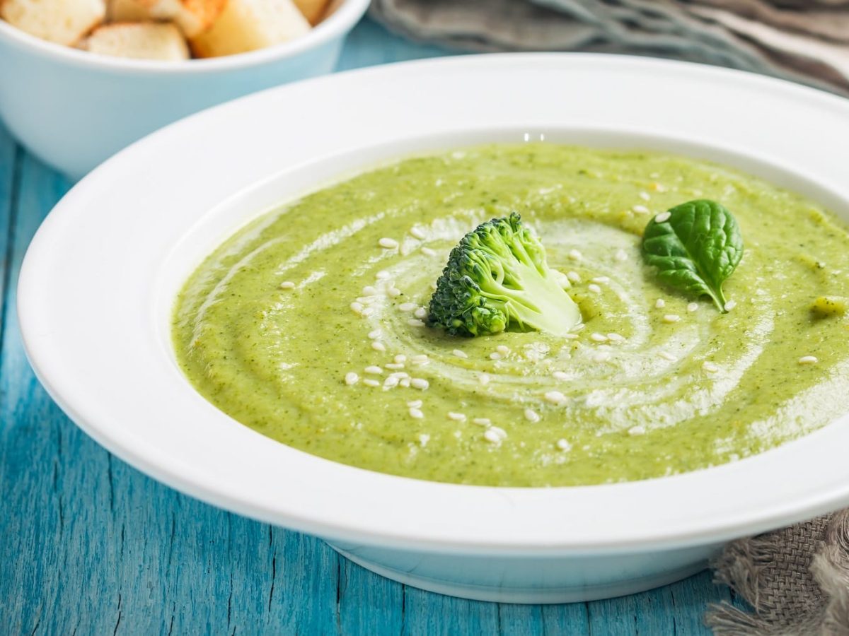 Ein weißer Teller mit der grünen Brokkoli-Cremesuppe auf einem bläulichen Holztisch.