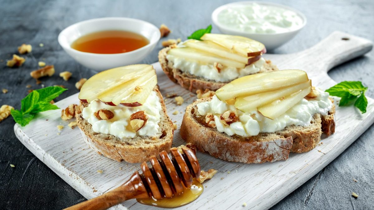 Drei Brotscheiben mit dem Hüttenkäse und Pfirsichspalten auf einem hellen Holzbrett.