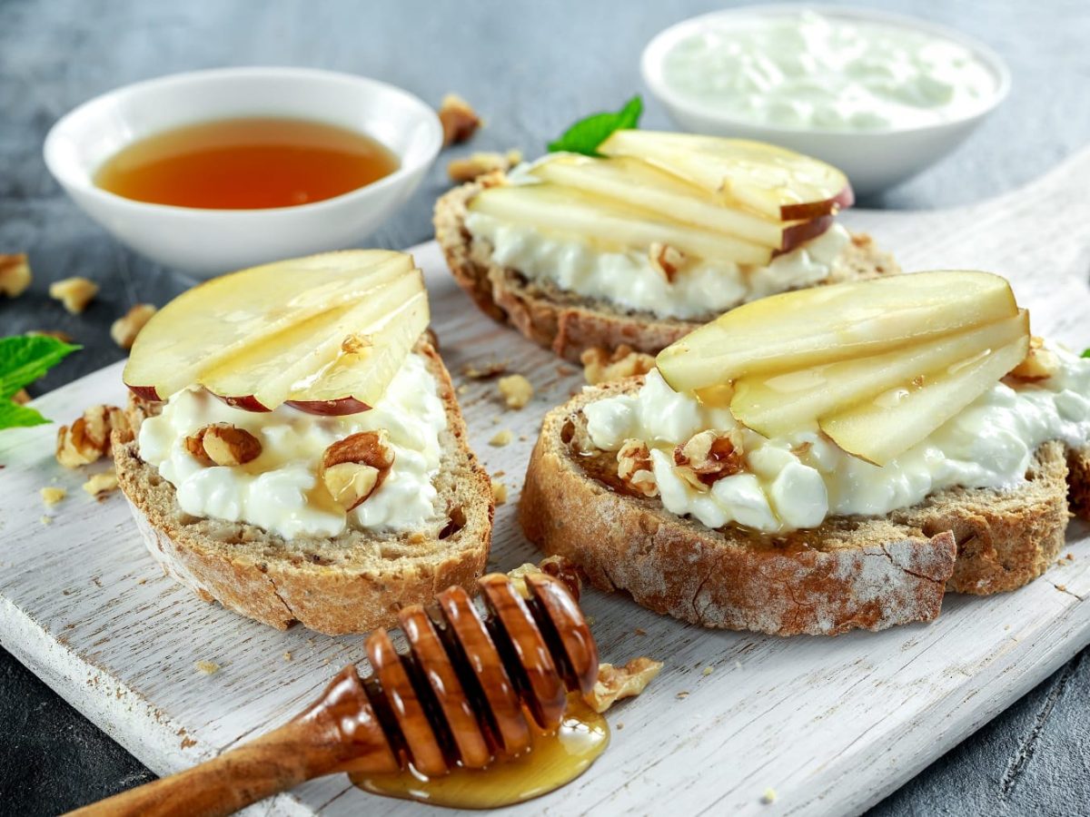 Drei Brotscheiben mit dem Hüttenkäse und Pfirsichspalten auf einem hellen Holzbrett.