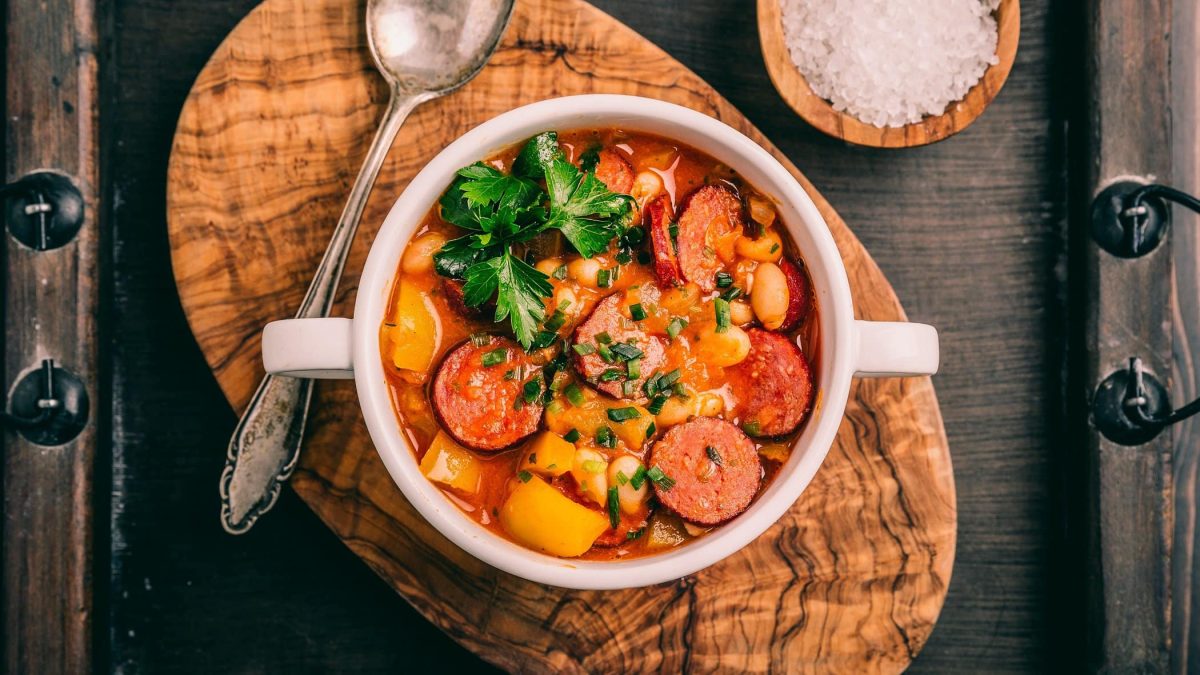 Draufsicht: Dunkler Untergrund. Auf dem liegt ein Holzbrettchen mit einem runden Schälchen Cabanossi-Stew mit Bohnen.