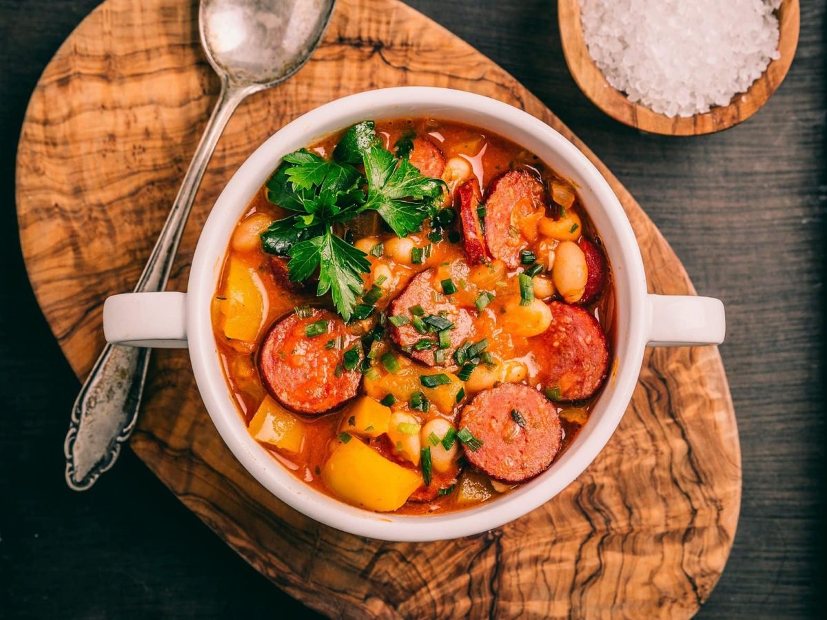 Draufsicht: Dunkler Untergrund. Auf dem liegt ein Holzbrettchen mit einem runden Schälchen Cabanossi-Stew mit Bohnen.