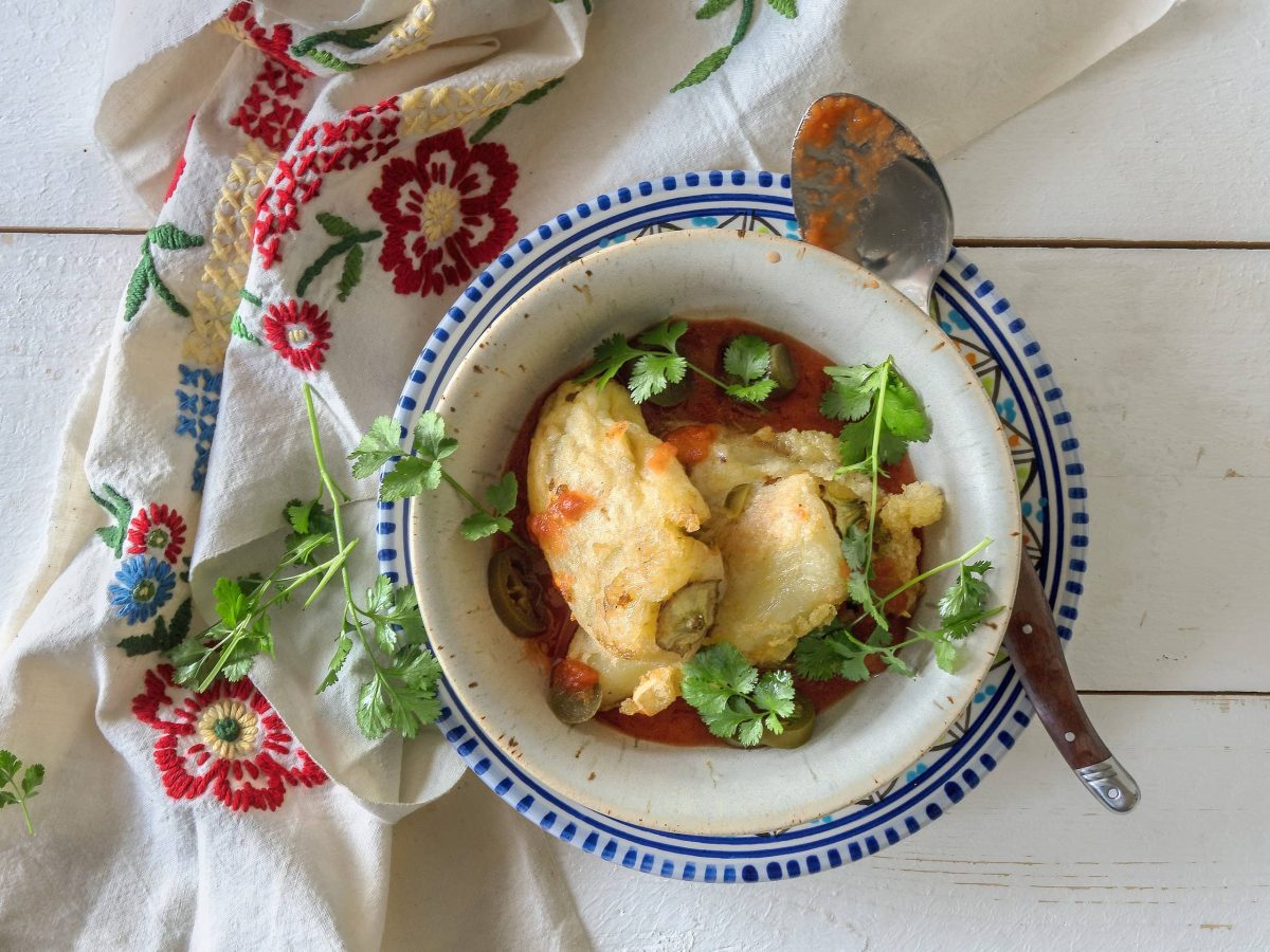 Chiles Rellenos con Queso, gefüllte Käsepaprika aus Mexiko, auf einem tiefen Teller, darin auch Tomatensauce, darauf ein Löffel und Koriander, daneben ein Geschirrtuch, alles Draufsicht.