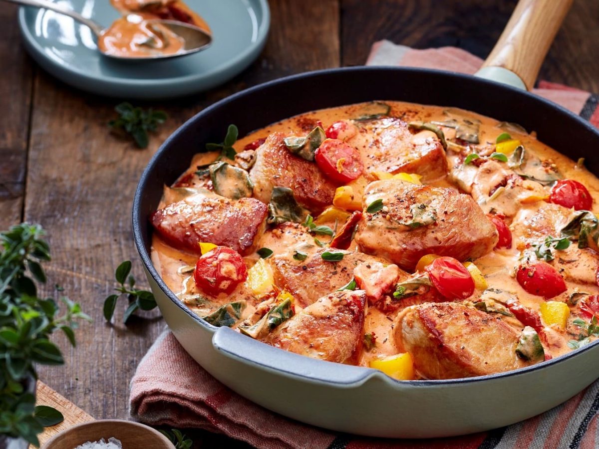 Eine schwarze Pfanne auf einem roten Geschirrtuch auf einem hölzernen Hintergrund. Die Pfanne ist gefüllt mit Pute in einer cremigen Tomatensauce. Links davon ein Kräuterstrauch.