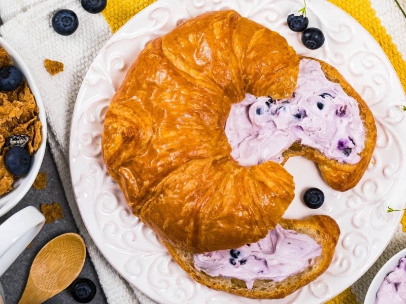 Draufsicht: Auf einem weißen Teller aus Porzellan liegt ein aufgeschnittenes Croissant mit Blaubeer-Thymian-Creme.