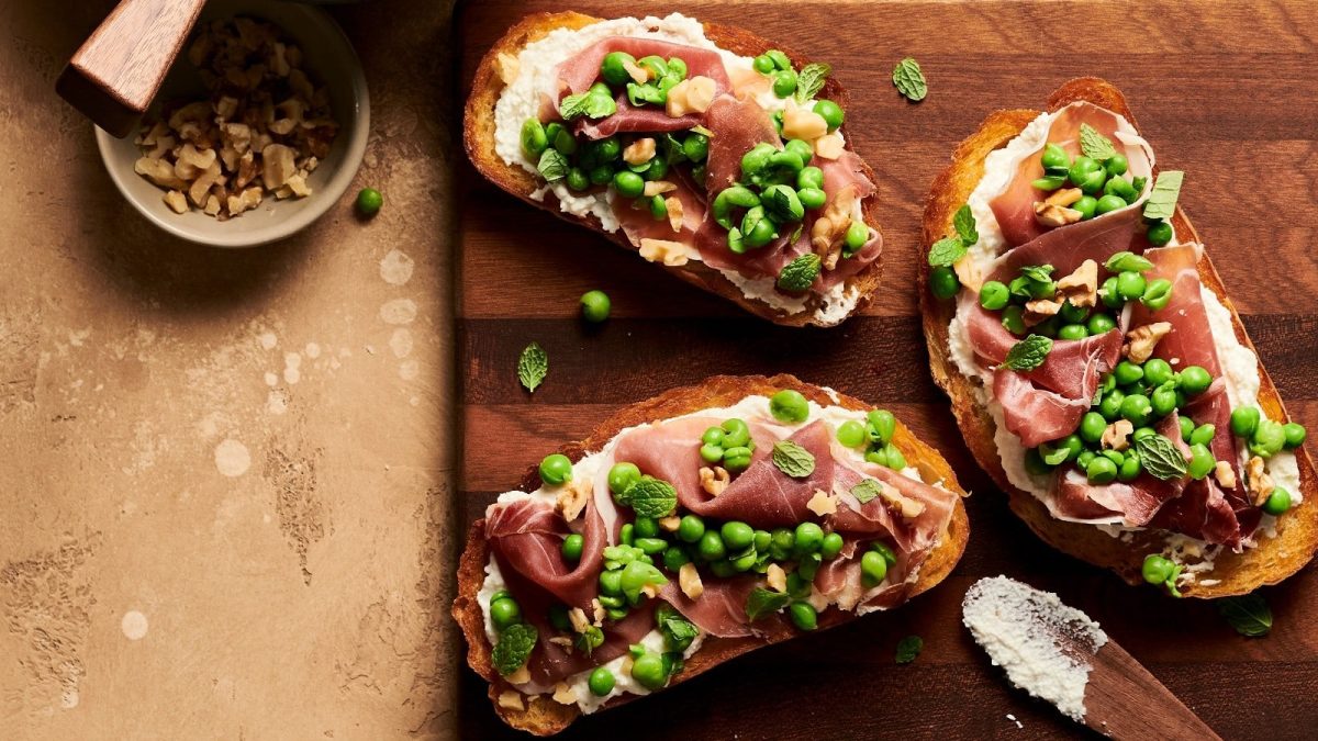 Ein Holzbrett mit Crostini mit Prosciutto und Erbsen auf einem Holztisch.