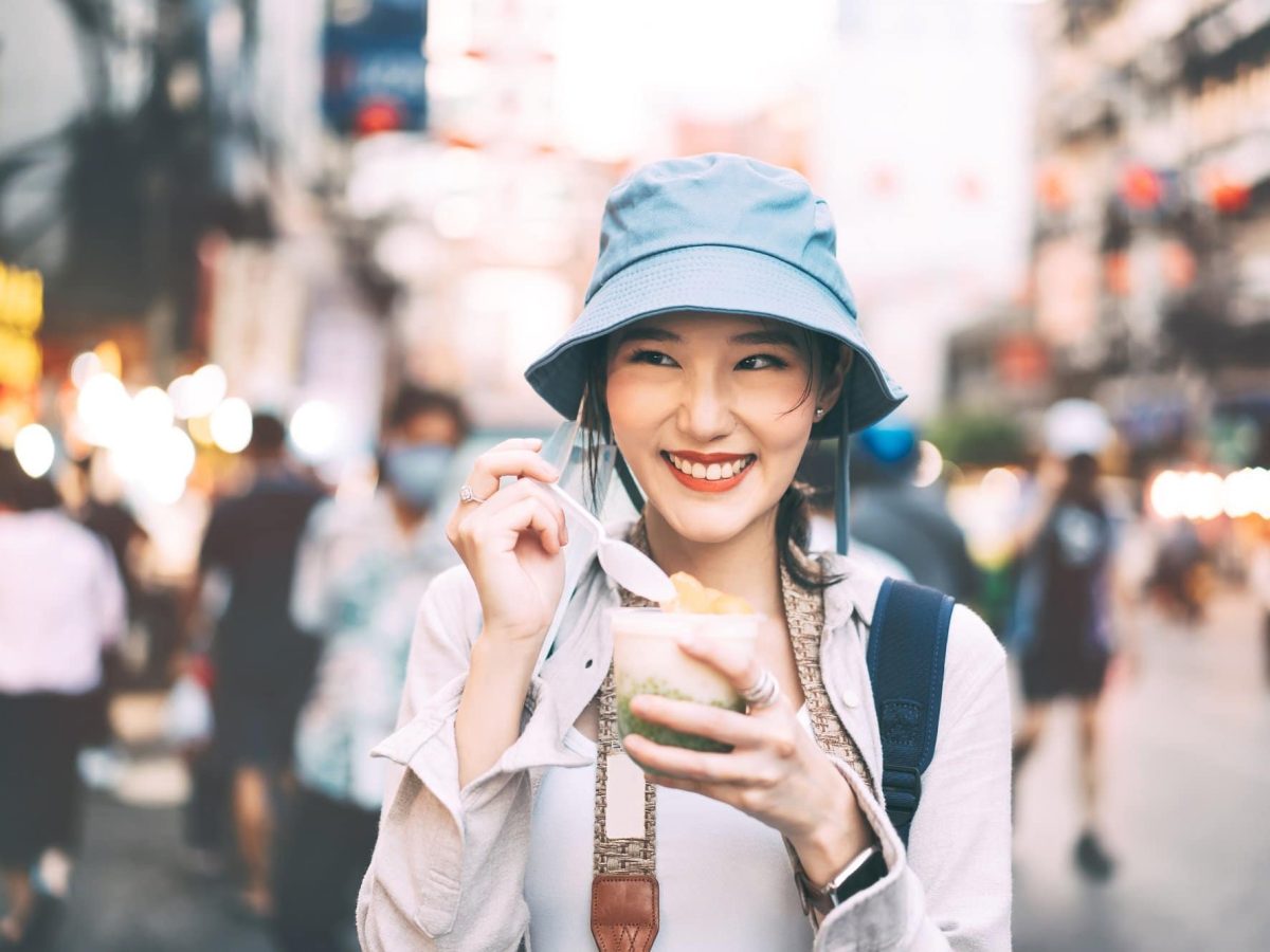 Vegane Städte: asiatische Touristin mit Hut, Rucksack und Eiscreme in der Hand, lächelnd, im Hintergrund unscharf eine Stadt.