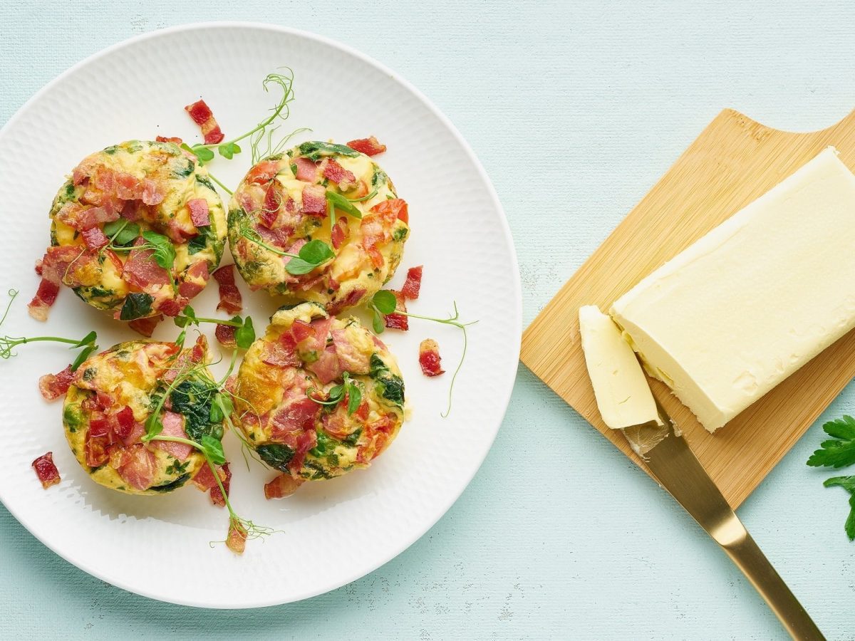 Draufsicht: Ein weißer Teller mit vier Eier-Muffins mit Speck und Spinat steht auf einem hellblauen Untergrund. Daneben liegen eine Butter auf einem Holzbrettchen, ein Küchenmesser und einige Blätter Petersilie.