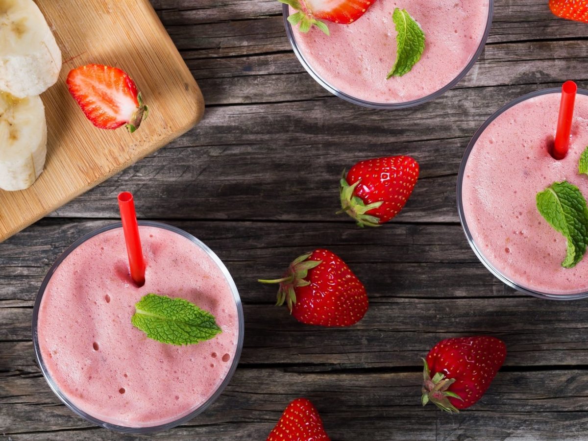 Mehrere Gläser mit dem Smoothie auf einem dunklen Holztisch mit Banane und Erdbeeren.