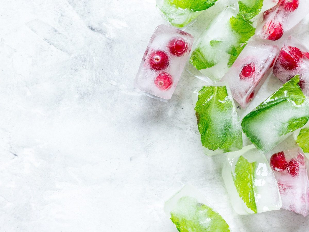 Draufsicht: Auf einer weißen Oberfläche aus Eis liegen Früchte-Eiswürfel und Kräuter-Eiswürfel.