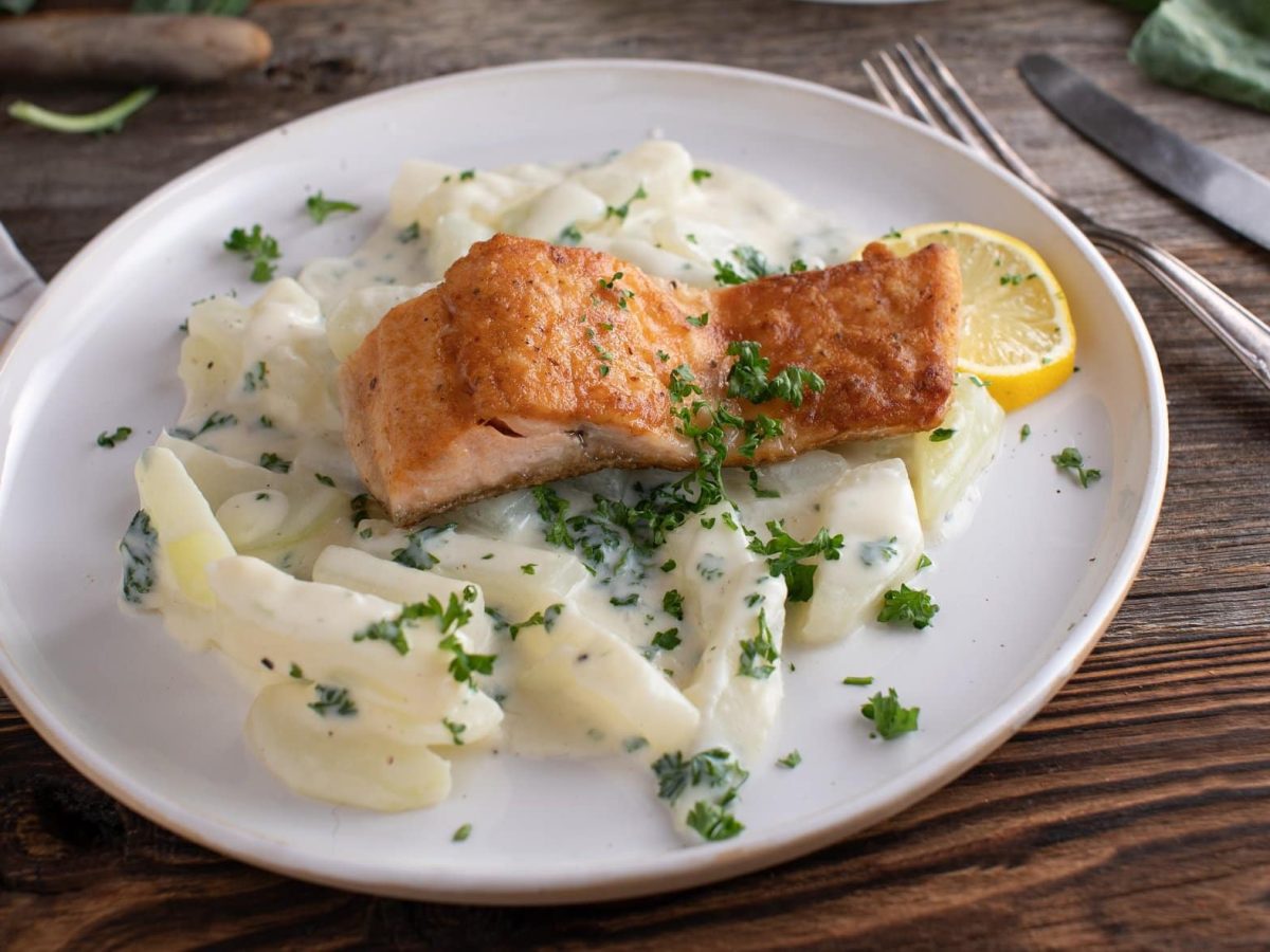 Draufsicht: Gebratener Lachs auf Kohlrabi auf einem weißen Teller liegt auf einem dunklen Holztisch. Daneben liegt Besteck.