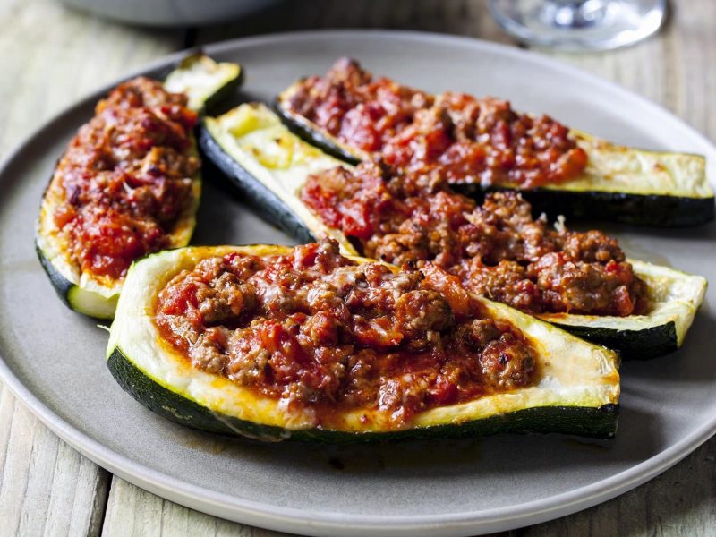 Frontalsicht: Auf einem Holztisch steht ein rustikaler Teller mit vier halben gefüllte Zucchini mit Lamm-Hackfleisch.