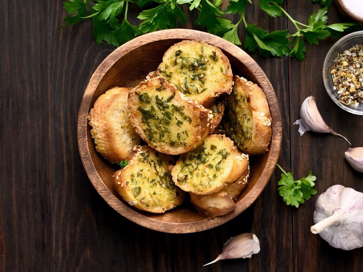 Draufsicht: Dunkler Untergrund, eine Korb mit geröstetem Knoblauchbrot mit Käse, frischer Knoblauch und frische Petersilie.