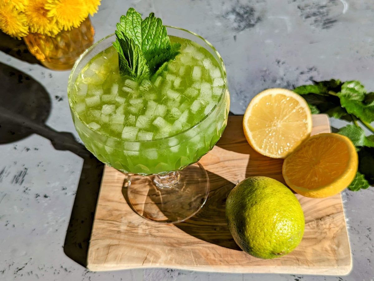 Der Green Limoni auf einem Holzbrett mit grauem Untergrund von schräg oben fotografiert. Im Hintergrund Zitronenmelisse und gelbe Blumen.